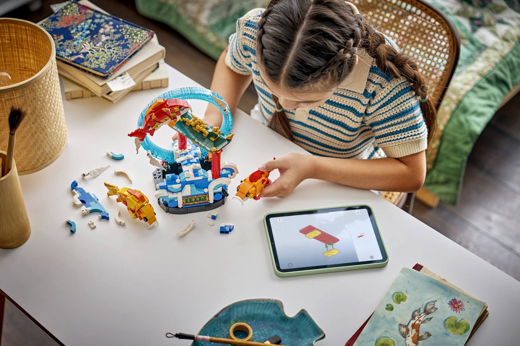 A girl holding a brick-built fish while using a tablet to build a LEGO® Chinese Festivals Prosperity Carp Leaping set.