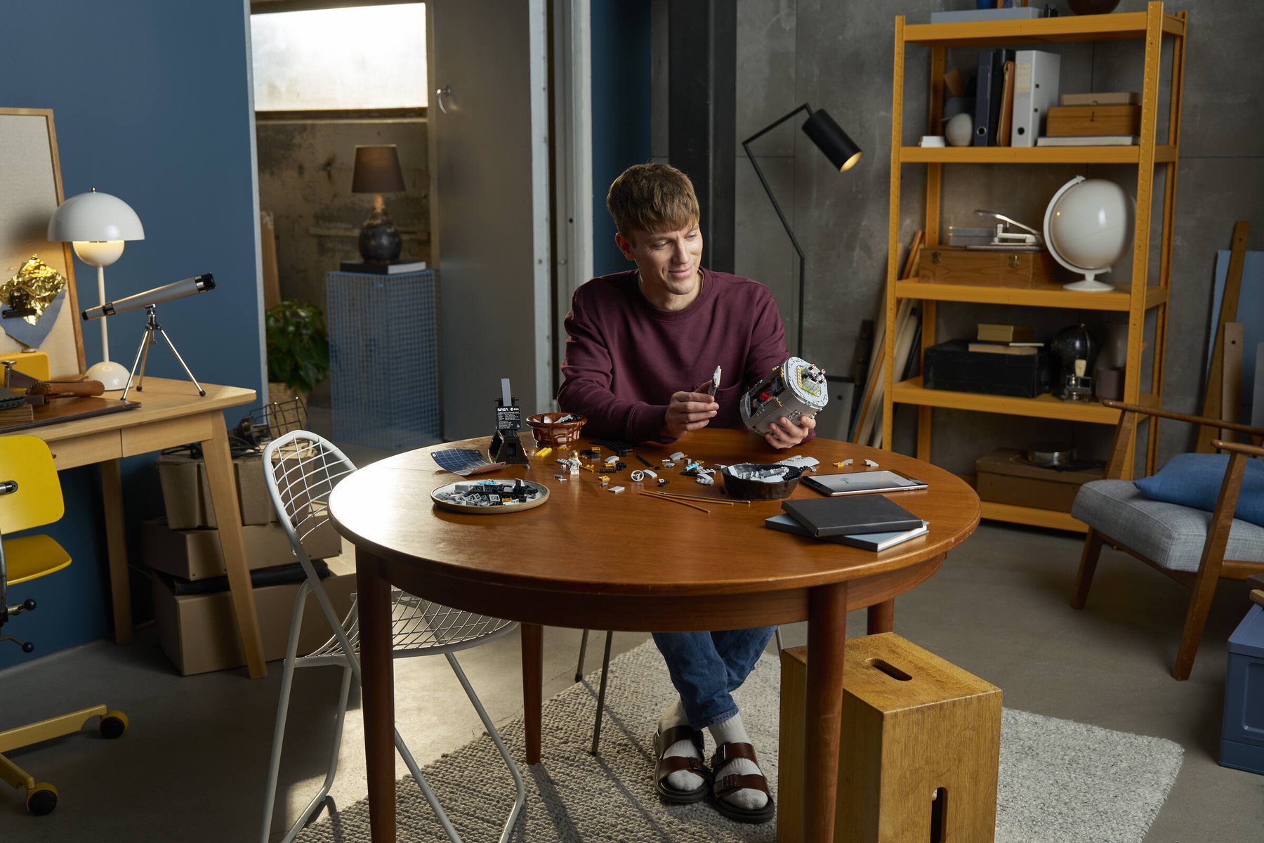 A man sitting at a table, surrounded by parts, using a tablet to build a LEGO® Icons Hubble Space Telescope set.