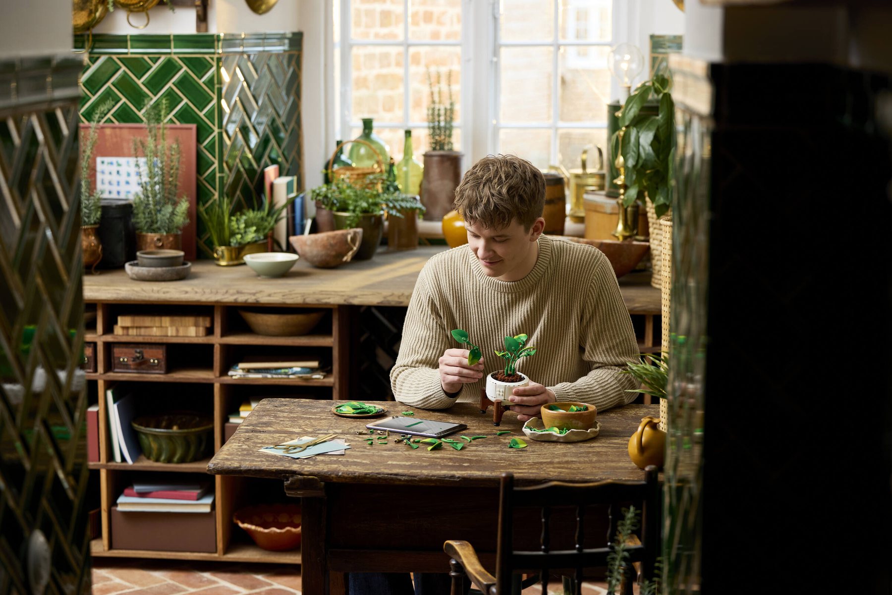 A man sitting at a desk holding a leaf piece while using a tablet to build a LEGO® Botanicals Hanging Golden Pothos set.