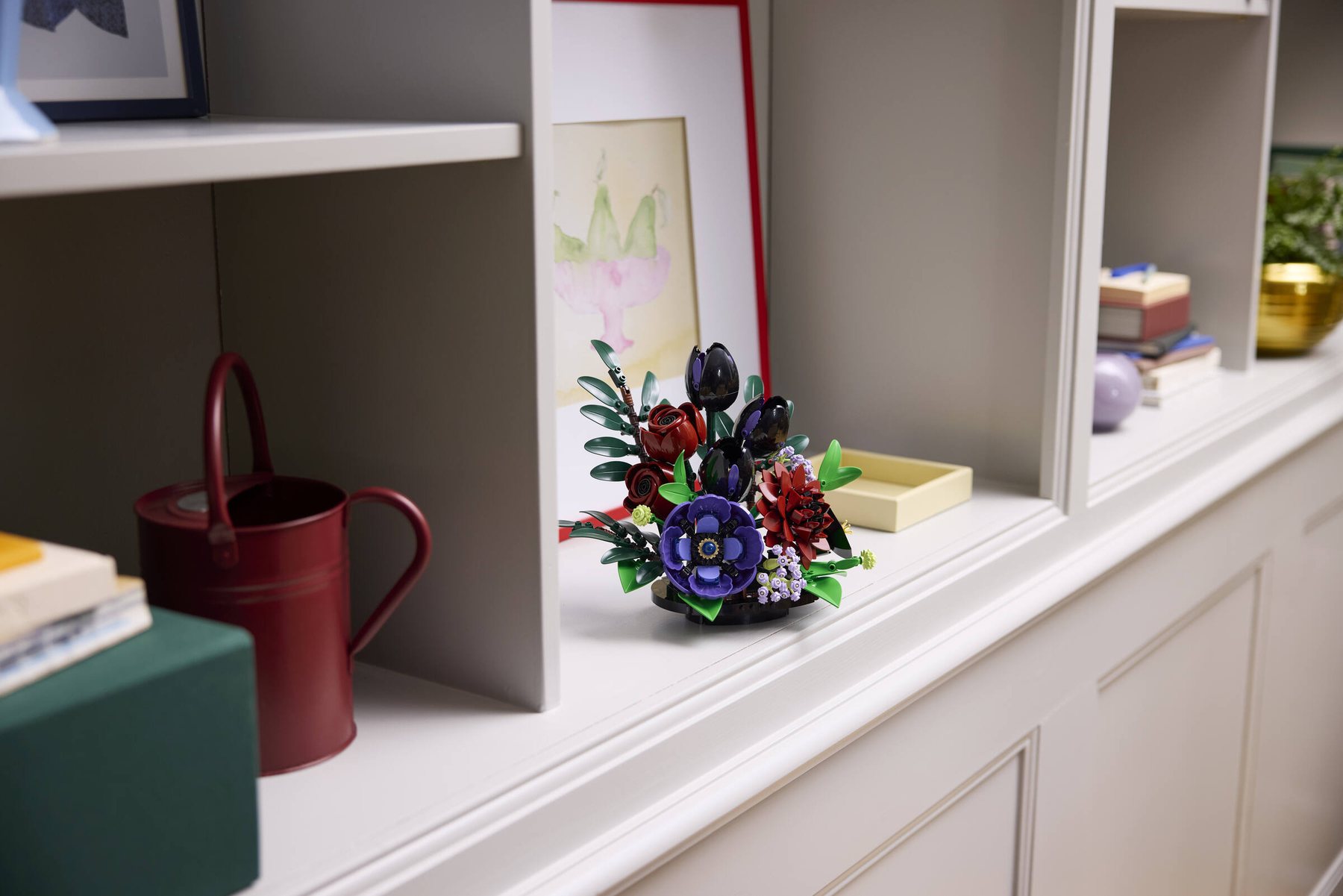 A woman sitting at a table adjusting a leaf in a LEGO® Botanicals Dark Flower Arrangement set. A LEGO® Botanicals Dark Flower Arrangement set displayed on a sideboard.