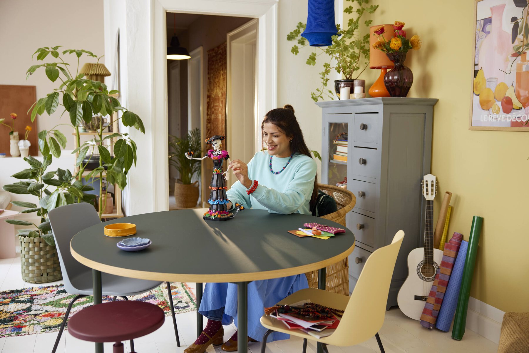 A woman sitting at a table adjusting the arm of the skeleton figure from a LEGO® Ideas La Catrina set.
